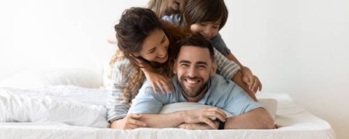 Portrait of happy young parents and small preschooler kids lying on white mattress in bedroom look at camera together, smiling family with little children relax on bed have fun, furniture concept